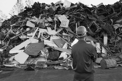 A man wearing a hard hat and a dark work jacket stands in front of a large pile of mixed waste materials, including broken furniture, plastic bins, metal scraps, and wood pieces, all piled high on a paved surface. The waste appears disorganized with various textures and colors, predominantly light and dark shades of plastic, wood, and metal. The background shows a clear outdoor setting without visible vegetation or structures, indicating an industrial or waste processing area, possibly related to garden clearance or rubbish removal services in Seven Kings. The man observes the waste pile, which is characteristic of an ongoing rubbish collection or disposal activity, typical of professional waste removal operations by companies like Garden Clearance Seven Kings. Lighting is natural, with even illumination across the scene, emphasizing the chaotic, unprocessed state of the debris, ready for sorting or transport.