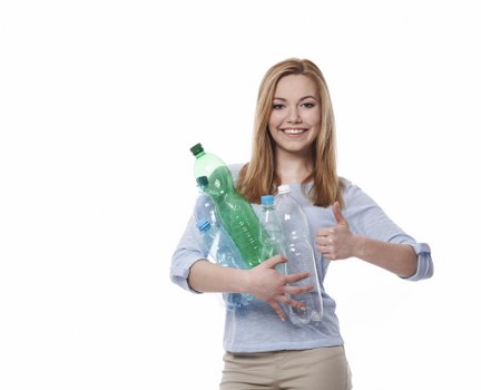 A young woman with shoulder-length blonde hair and a friendly smile holds multiple clear plastic bottles, including one green-topped bottle, against her chest. She is wearing a light blue long-sleeved shirt and beige trousers, standing in a neutral setting. Her right arm is crossed over her body, supporting the bottles, while her left hand is giving a thumbs-up gesture. The bottles are different sizes and shapes, typical of household recyclables. The background is plain white, emphasizing her and the bottles she is holding. This image depicts a positive attitude towards recycling and rubbish collection, aligning with services provided by Garden Clearance Seven Kings. The visual focus on plastic waste materials supports local waste removal and recycling initiatives in the Seven Kings area, indirectly referencing the importance of responsible rubbish disposal practices. The clear, natural lighting highlights the textures and colours of the bottles, and her casual attire suggests a community-oriented approach to rubbish management services.