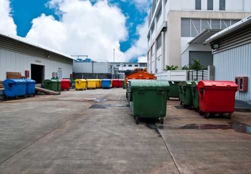 A wide view of an outdoor industrial area showing a concrete surface with several large wheelie bins in various colours including green, red, yellow, grey, and blue, arranged along the edges of the space. The bins are made of plastic with textured surfaces, and some have lids closed while others are open. In the background, there are two metal-sided buildings, one on the left with a small gap at the bottom showing a glimpse of a door or window, and a taller white building with multiple floors and windows on the right. Behind the bins, a flat-roofed industrial structure with blue piping and vents is visible. The sky above is partly cloudy with patches of blue, indicating daytime, and the scene suggests a waste collection or rubbish removal area typical of commercial premises in the Seven Kings area, which is within the vicinity of London. The overall environment appears clean and organized, overseen by Garden Clearance Seven Kings, specialists in waste removal services in the local region.